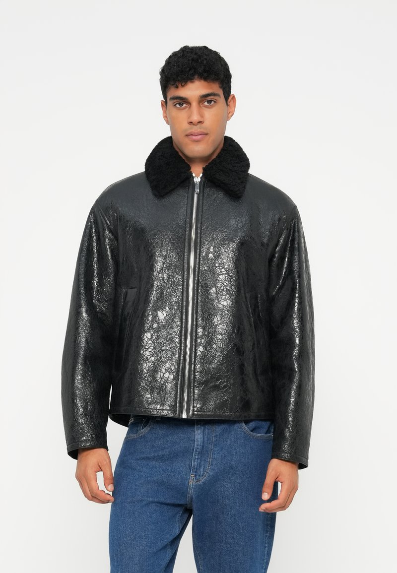 Jaqueta de couro sintético preta com acabamento brilhante, apresentando um colarinho felpudo preto e fecho frontal com zipper, combinada com jeans de ganga azul.