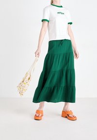 Valkoinen t-paita vihreillä yksityiskohdilla ja logolla, yhdistettynä pitkään vihreään kerrokselliseen hameeseen ja oransseihin korkokenkiin. Luonnonmateriaalista valmistettu laukku keltaisilla yksityiskohdilla.