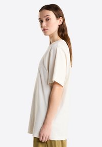 Mujer joven con cabello largo y castaño, lleva una camiseta grande de color blanco roto y pantalones oliva, está de pie de perfil contra un fondo claro uniforme.