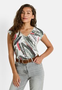 Camiseta de manga corta con escote en V en blanco con un patrón abstracto multicolor, que presenta acentos en verde, rojo y dorado, combinada con jeans grises y un cinturón marrón.