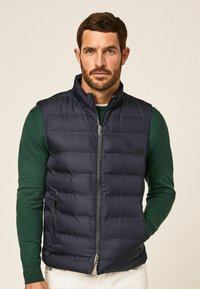 Marineblaues Steppweste mit Reißverschluss, verfügt über einen hohen Kragen, Seitentaschen und ein gestepptes Design, getragen über einem grünen Kleidungsstück.