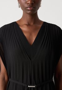 Black pleated dress with a deep V-neckline, short sleeves, and a fitted waist belt featuring a metallic accent. Smooth texture and flow.