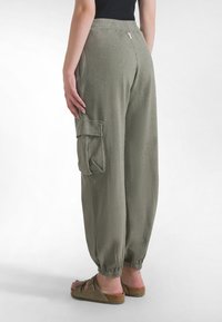Spodnie cargo w oliwkowej zieleni o luźnym kroju, z elastycznym pasem, ściągaczami przy nogawkach i ozdobnym bocznym kieszeniem. Materiał wydaje się lekki i teksturowany.