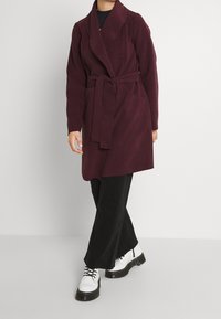 Abrigo burdeos de manga larga con cinturón, gran collar y dos bolsillos delanteros, combinado con pantalones negros y botas blancas con cordones.