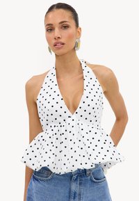 Femme portant un haut dos-nu blanc à pois noirs avec un ourlet à volants, associé à un jean bleu et de grandes boucles d'oreilles géométriques.