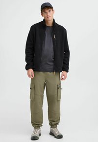 Chaqueta de forro polar negra con cuello alto, camiseta gris debajo, pantalones cargo oliva con bolsillos y botas de senderismo grises. Gorra sencilla en la cabeza.