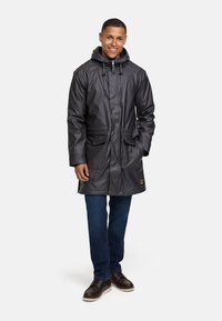 Chaqueta impermeable gris con capucha, con cordones, dos bolsillos delanteros y un corte relajado, llevada sobre unos vaqueros azules y botas marrones.