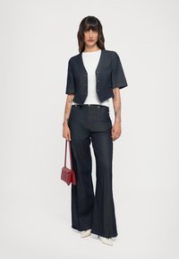 Giacca in denim scuro cropped con maniche corte e bottoni, abbinata a pantaloni in denim scuro a campana. Camicia bianca sotto, mentre tiene una borsa rossa.