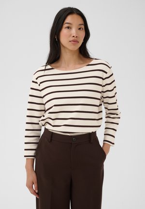 Mujer con cabello largo y negro, que lleva una camisa blanca de manga larga con rayas negras y pantalones de talle alto de color marrón oscuro, mano en el bolsillo.