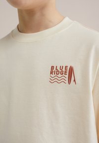 Kremowa koszulka z miękkiej bawełny, z czerwonym grafiką przedstawiającą napisy "BLUE RIDGE", fale i zarys deski surfingowej.