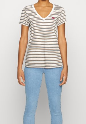 Person iført en hvid t-shirt med korte ærmer, V-hals med tynde horisontale sorte og brune striber og lyseblå jeans.