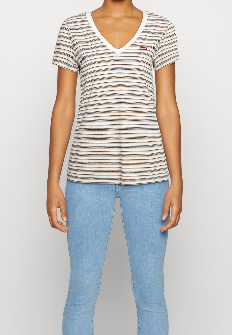 Personne portant un t-shirt blanc à manches courtes et col en V avec de fines rayures horizontales noires et marron et un jean bleu clair.