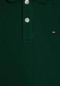 Polo de color verde oscuro hecho de tela texturizada, con una abertura de tres botones y un pequeño logotipo bordado en rojo, blanco y azul.
