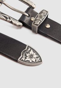Cinturón de cuero negro con una hebilla de metal grabada en plata con un diseño floral y una punta; presenta una superficie texturizada y un detallado ornamental.