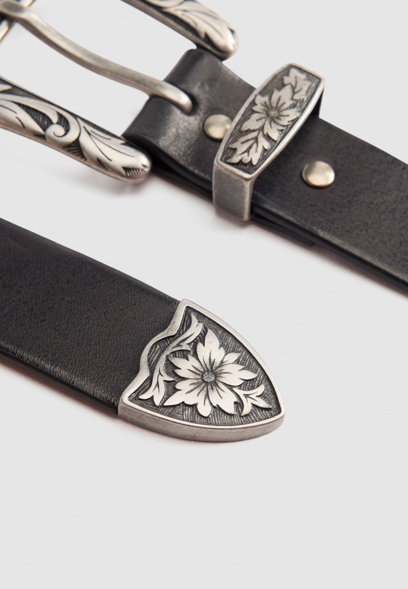 Cinturón de cuero negro con una hebilla de metal grabada en plata con un diseño floral y una punta; presenta una superficie texturizada y un detallado ornamental.