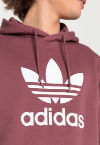 Burgundy hoodie med en stor vit Adidas-logotyp, inklusive en trefloddesign med tre streck över bröstet och dragsnodd vid huvan.