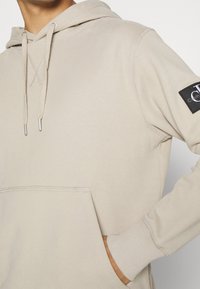 Sudadera de algodón beige con capucha ajustable, bolsillo tipo canguro y puños acanalados. Presenta un parche de logo negro en la manga izquierda.