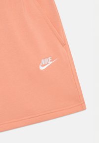 Ferskenfargede Nike-shorts laget av myk stoff, med to sidelommer og en hvit brodert logo nederst til venstre.