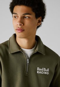 Sudadera de cremallera verde oliva con cuello, presenta "Red Bull RACING" bordado en blanco. Textura suave, diseño minimalista con cremallera plateada.