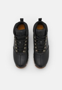 Pár černých kožených turistických bot s kovovými očky a hnědými podrážkami, zobrazený zhora na jednoduchém bílém pozadí.