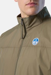 Giacca verde oliva con collo alto, chiusura con zip e un logo circolare blu e bianco. Il tessuto sembra liscio e leggero.
