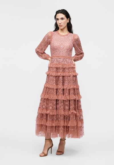 Γυναίκα στέκεται με τα χέρια στη μέση, φοράει ένα μακρύ, σκουρόροζ δαντελωτό φόρεμα με στρωτά βολάν και διάφανα φουσκωτά μανίκια.