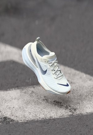 Chaussure de course Nike blanche avec swoosh bleu, en l’air au-dessus d’un bitume gris avec une bande blanche, montrant la semelle et les lacets.
