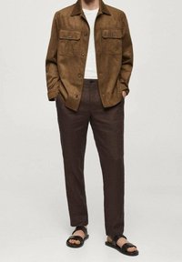 Veste en daim marron avec deux poches poitrine, chemise blanche, pantalon marron et sandales noires. La tenue présente une coupe décontractée et un design casual.