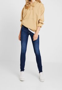 Sudadera beige con bolsillo frontal y capucha con cordón, combinada con vaqueros ajustados de color azul oscuro y zapatillas blancas. Textura de tejido suave en toda la prenda.