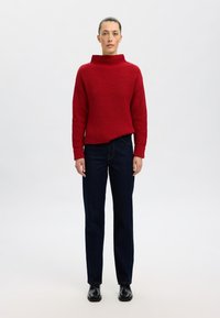 Roter ribbeliger Strickpullover mit hohem Kragen, kombiniert mit geraden Jeans aus dunklem Denim. Das Model steht vor einem schlichten Hintergrund. Schwarze Stiefel.