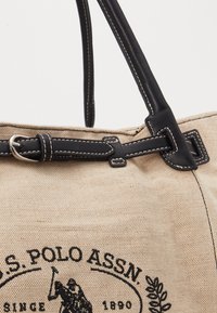 Beige canvasväska med svarta läderremmar och spänne. Har vit sömnad och en tryckt logotyp av en polo-spelare.