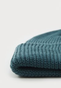 Teal knitted beanie with a ribbed texture, folded edge, and soft, wool-like appearance. Evenly spaced stitching creates a uniform pattern.