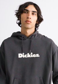 Hanorac gri din material moale, cu un buzunar frontal și șnururi. Logo-ul alb mare "Dickies" pe piept.