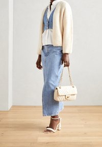 Žena stojí oblečená v lehkém kardiganu, s vrstveným denimovým outfitem a na vysokých podpatcích, drží béžovou kabelku na neutrálním pozadí.