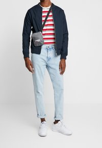 Uomo che indossa una giacca bomber blu navy, una maglietta a righe rosse e bianche, jeans chiari arrotolati, sneakers bianche e una borsa a tracolla nera.