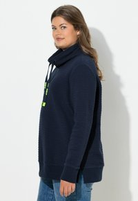 Sudadera azul marino con tejido texturizado, cuello alto y cordones ajustables en verde neón. Puños y bajo acanalados.