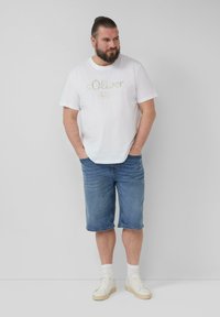 Weißes Baumwoll-T-Shirt mit goldenem Logodruck, kombiniert mit knielangen blauen Jeansshorts. Tragen mit weißen Sneakers und Knöchelsocken.