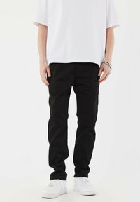 Sorte tapered bukser lavet af bomuldsblanding med en glat tekstur og forlommer, parret med hvide sneakers. Hvid t-shirt synlig ovenfor.