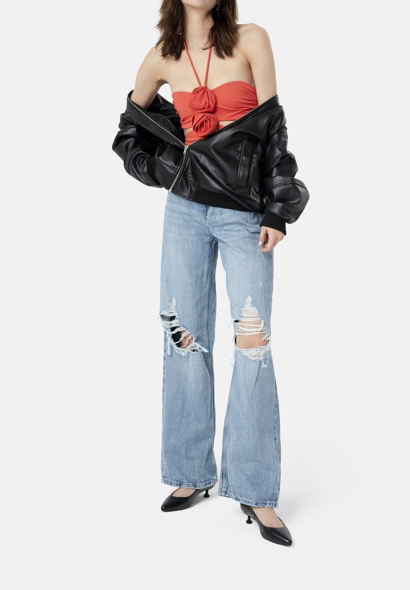 Femme portant un crop top rouge à col halter avec des détails floraux, une veste en cuir noire oversize, un jean bleu clair déchiré et des talons noirs pointus.