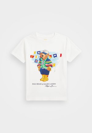 Camiseta blanca de algodón con una ilustración colorida de un oso con chaqueta a rayas, camisa verde y botas amarillas, con banderas náuticas de fondo.