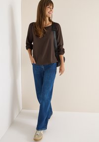 Top lungo marrone a maniche lunghe con vestibilità morbida, abbinato a jeans blu e sneakers. Gli accessori includono una borsa a tracolla nera. Sfondo chiaro.