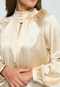 Blouse en soie crème avec un col haut, un détail en trou de serrure et des manches bouffantes. Texture lisse et accents froncés le long de l'encolure et des poignets.