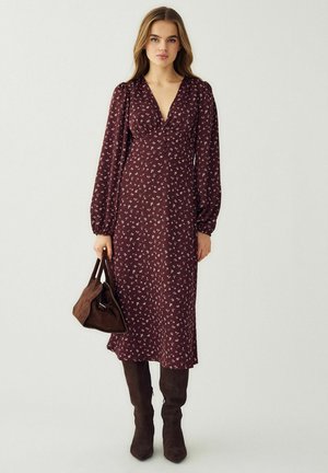 Ung kvinde iført en langærmet, bordeaux floral midi-kjole, holder en brun håndtaske og har mørkebrune knæhøje støvler på.