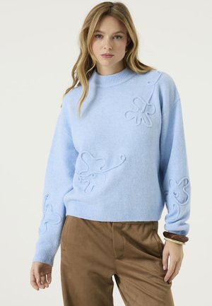 Vrouw met een lichtblauwe trui met verhoogde abstracte bloemenpatronen, een bruine broek en een armband, staand tegen een effen achtergrond.