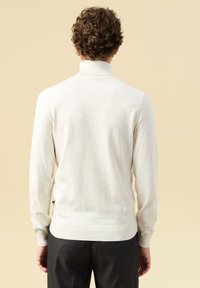 Jasno beżowy sweter z wysokim kołnierzem, prążkowanymi mankietami i dołem. Miękka tekstura, dopasowany krój oraz minimalny detal z logo przy dole.