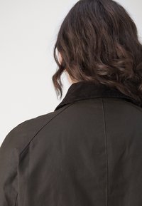 Personne avec de longs cheveux bruns ondulés portant une veste marron foncé avec un col en velours côtelé, vue de dos sur un fond clair.
