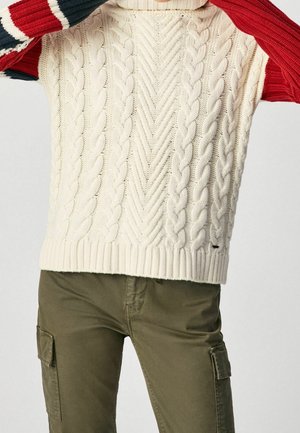 Personne portant un pull sans manches crème en maille torsadée sur une chemise rayée rouge, blanche et noire à manches longues, avec un pantalon cargo vert olive.