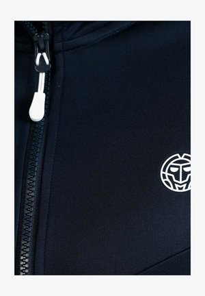 Close-up van zwarte stof met een wit plastic ritslipje en een wit cirkelvormig logo met een gestileerd gezicht.