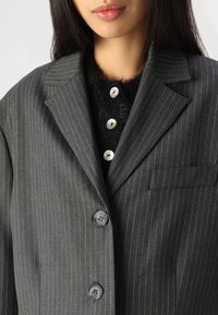 Grå stribet blazer med notch lapels, to knapper og en brystlomme. Båret over en tekstureret sort sweater med runde knapper.