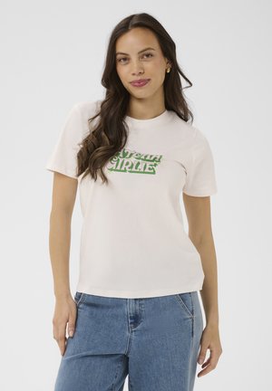 Femme aux longs cheveux bruns portant un t-shirt blanc avec un texte vert et un jean bleu, se tenant devant un fond clair uni.